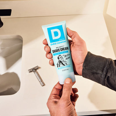 Hand holding Duke Cannon Shave Cream tube with a razor on a bathroom counter.
